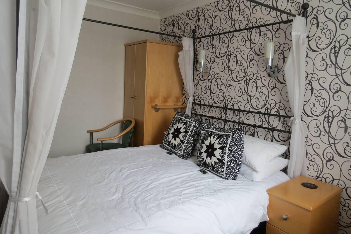 A bed with a canopy, a wooden wardrobe, a bedside table, and a wall with a black and white patterned wallpaper.