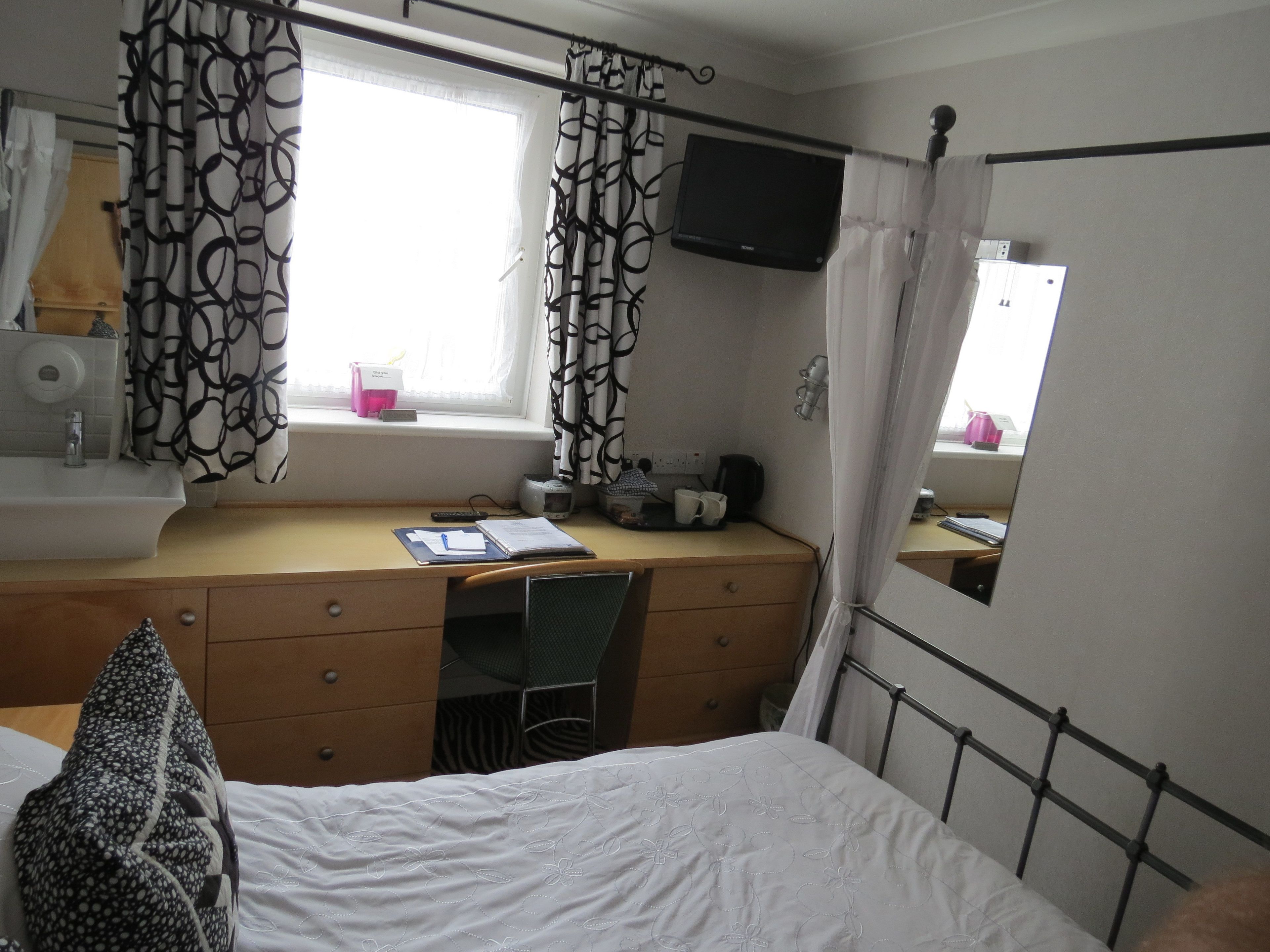 Un dormitorio con cama, escritorio, espejo, televisor y ventana con cortinas.