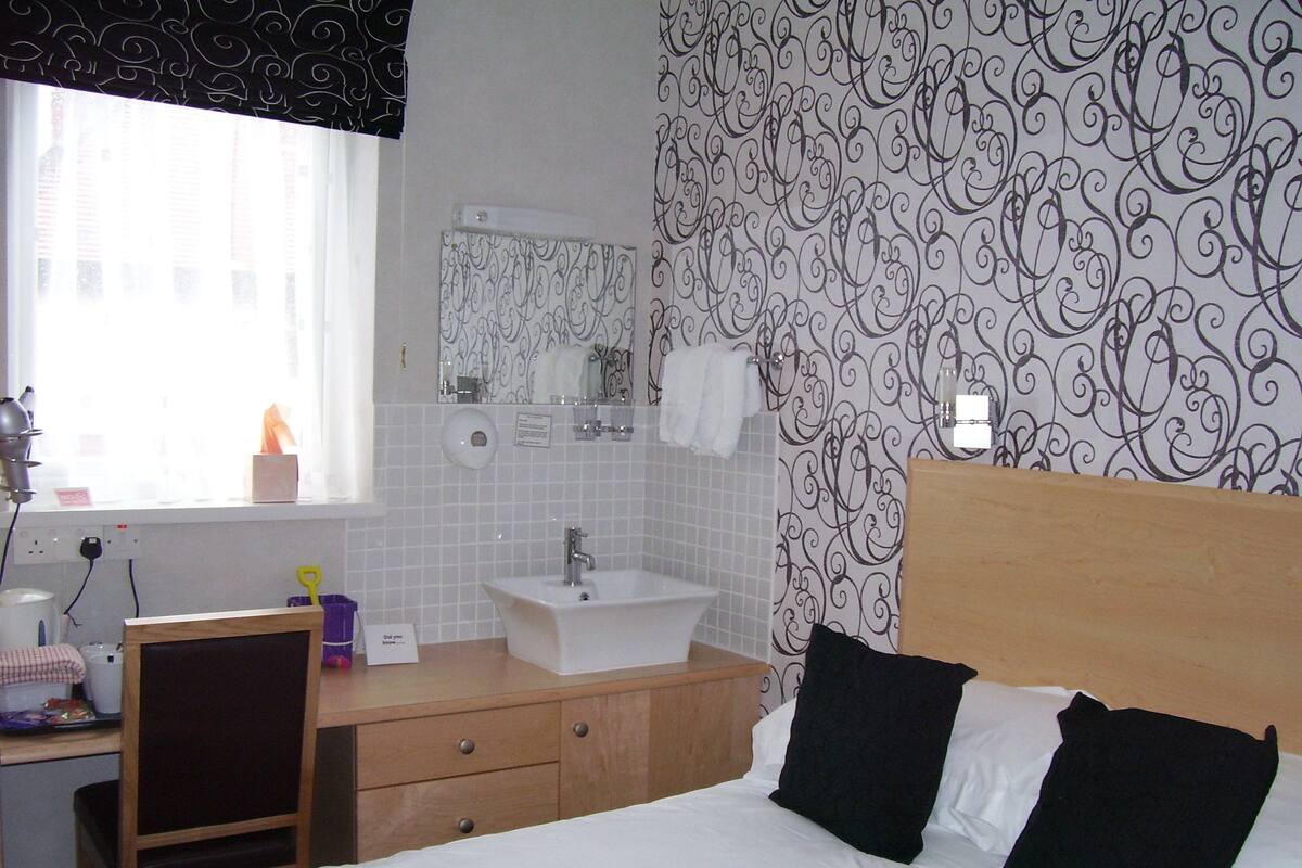 A hotel room with a bed, a wooden desk, a sink, and a mirror.