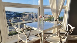 A dining area with a glass table and chairs, a view of the city and sea, and a window with white curtains. - Harbour View Guest House (St Ives)