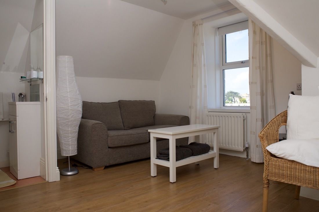 Una habitación pequeña y acogedora con un sofá gris, una mesa blanca y una silla de mimbre. Se ve un radiador debajo de una ventana, y una lámpara blanca está al lado del sofá.
