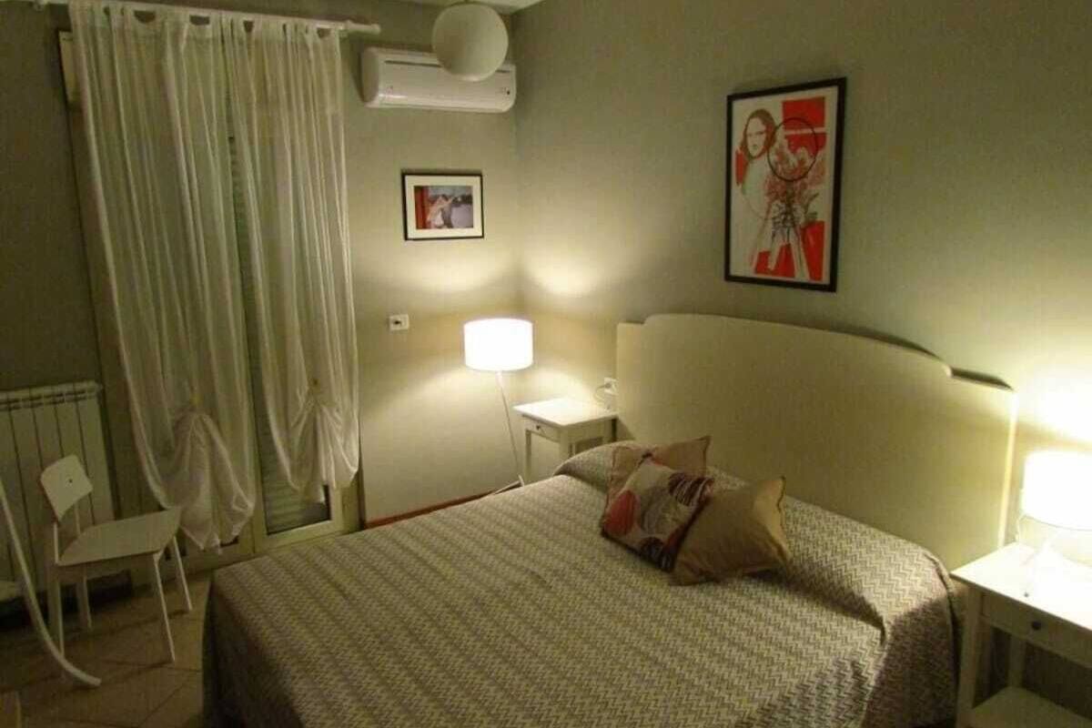 Hotellihuone, jossa on sänky, tuoli, yöpöytä, lamppu ja taulu seinällä.