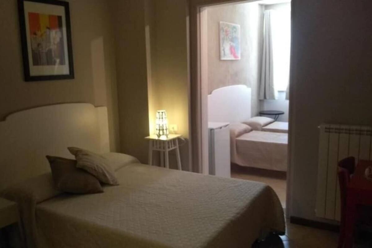 Hotellihuone, jossa on sänky, yöpöytä, lamppu, tuoli ja ovi, joka johtaa toiseen huoneeseen.