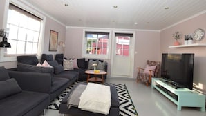 A modern living room with a grey sofa, a small round table, a TV on a stand, and a large window with a view of buildings. - Solferie Holiday Home Jolleveien (Kristiansand)