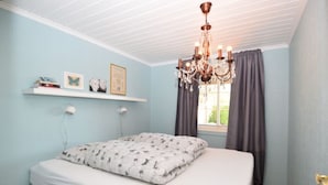 A bedroom with a bed, a chandelier, a window with curtains, and a shelf with books and pictures. - Solferie Holiday Home Jolleveien (Kristiansand)