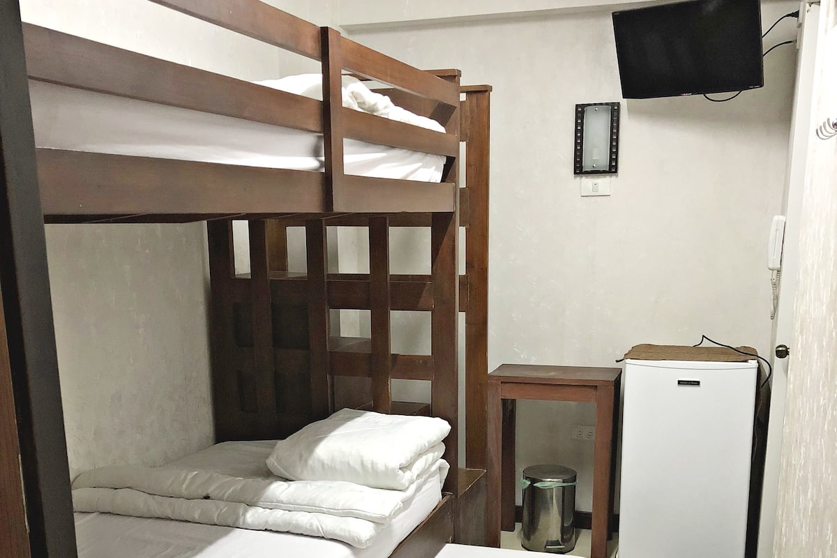 A bunk bed with white bedding, a small table with a lamp, a refrigerator, and a mounted TV.