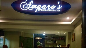 A restaurant interior with a sign reading "Amparo Kitchen, Coffee & Tea". There are wooden chairs and tables, and a coffee cup in the foreground.