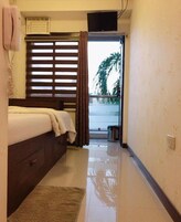 Habitación de hotel con cama, una mesita, ventana con persianas, televisor montado en la pared y vistas al exterior.
