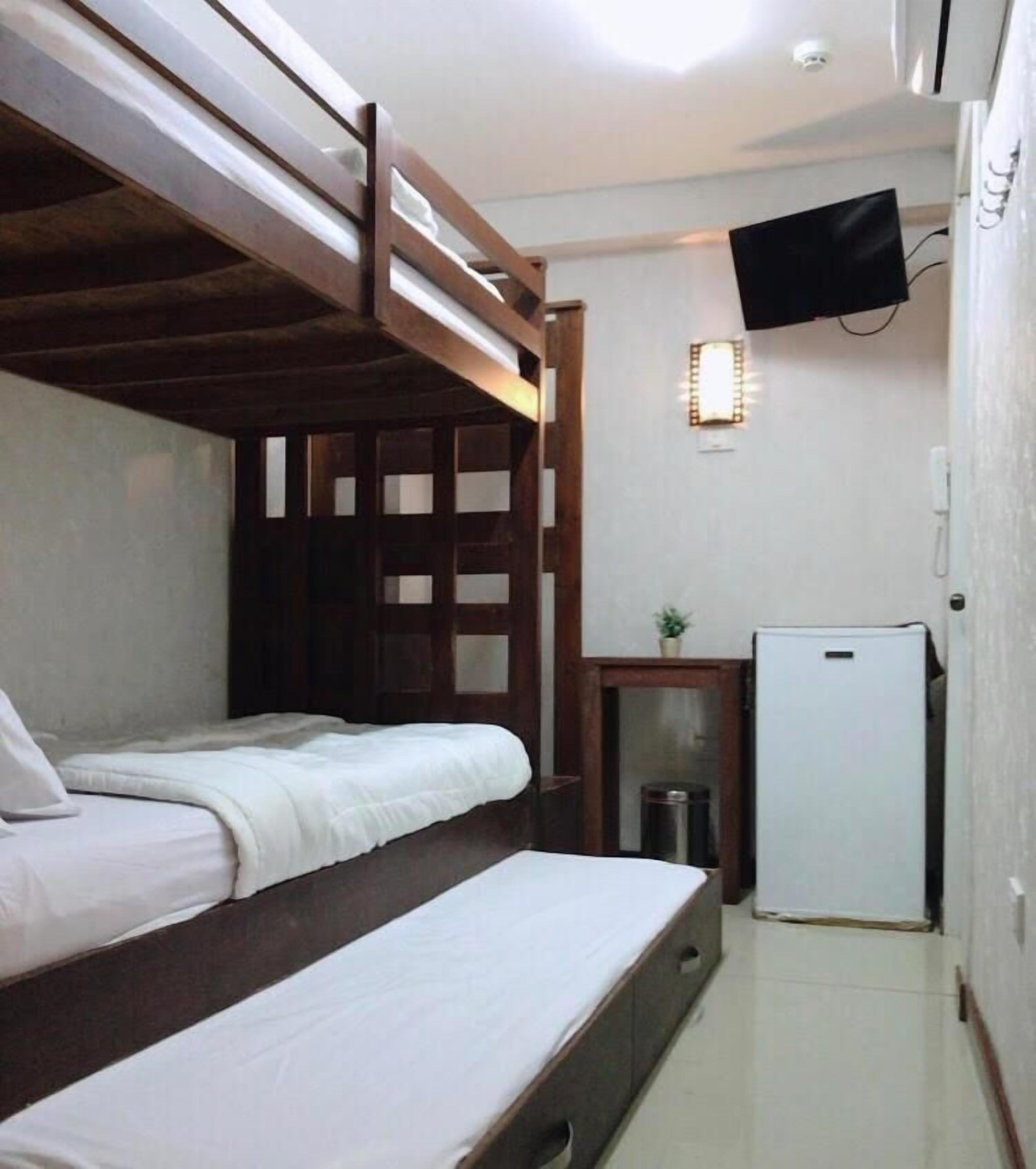 A compact room with two bunk beds, a small white refrigerator, a television mounted on the wall, and a trash bin.