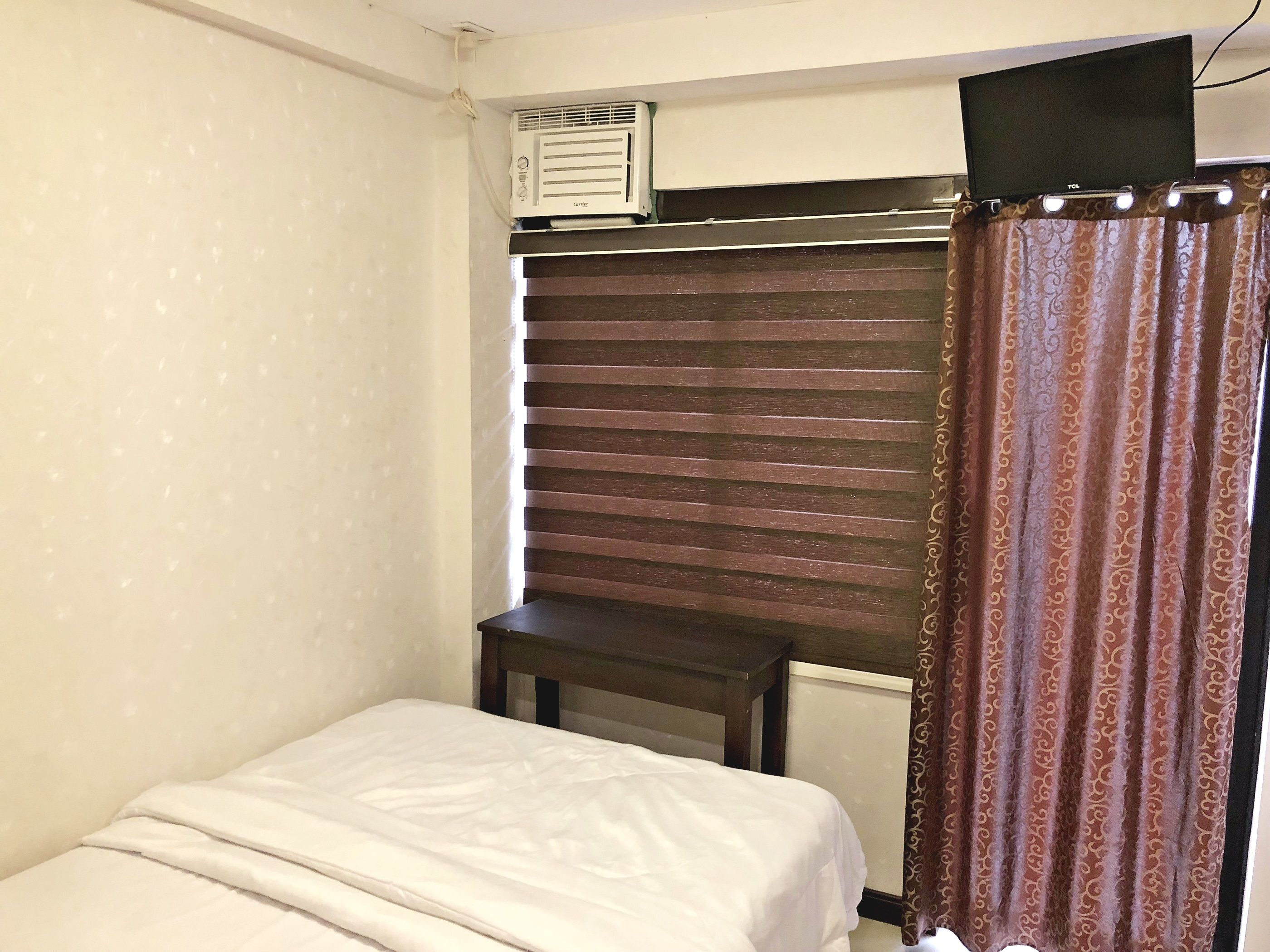 Una habitación pequeña y funcional con cama, una mesita de madera, cortina y aire acondicionado.
