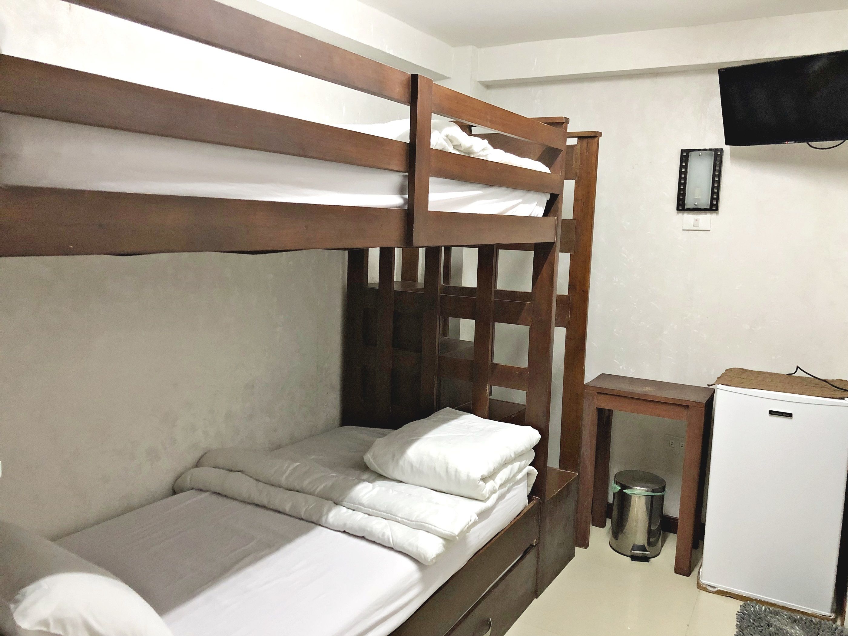 A bunk bed with white bedding, a small wooden desk, a trash can, and a white refrigerator.