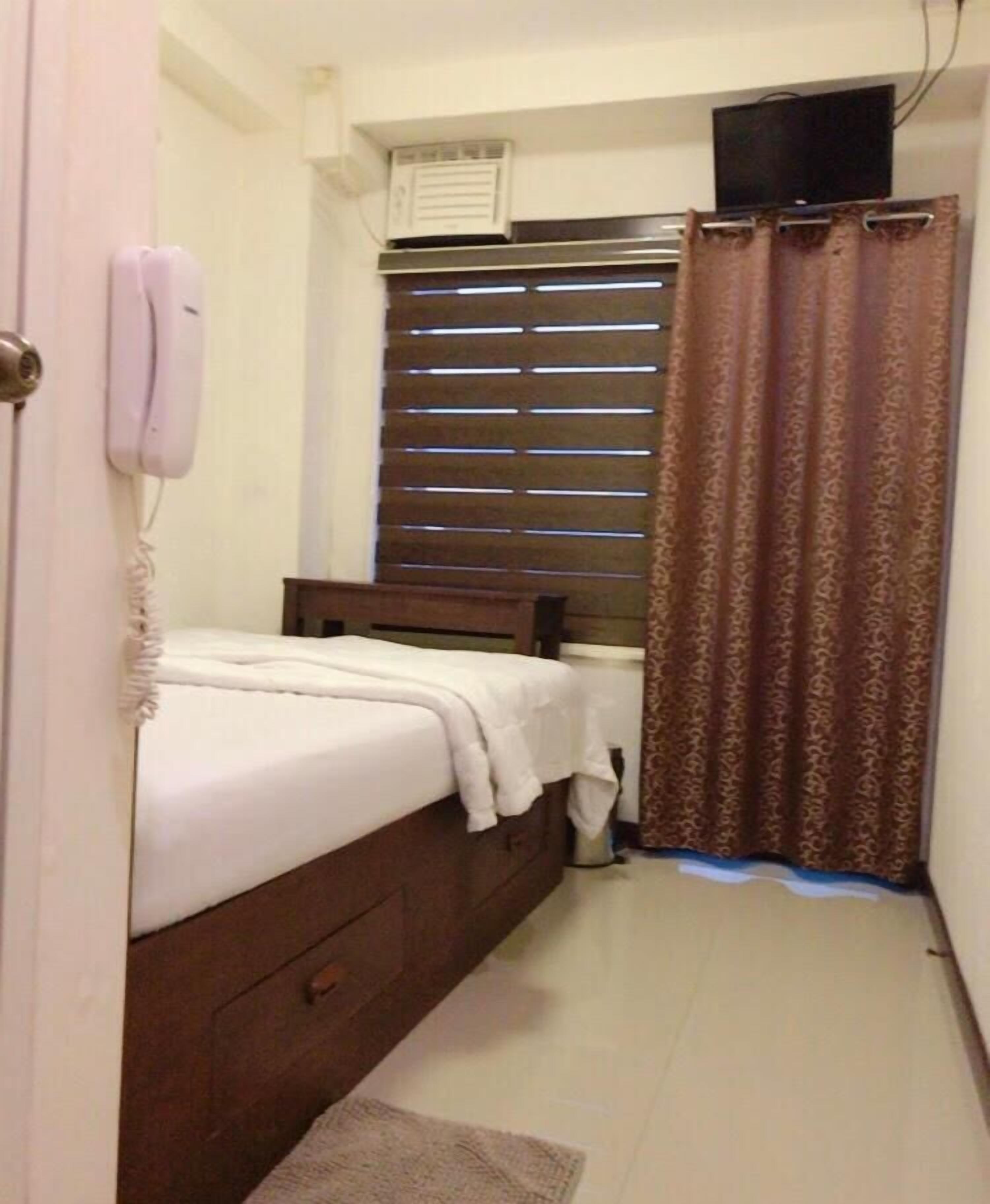 Un dormitorio pequeño y ordenado con una cama individual, un teléfono en la mesita de noche, una ventana con persianas y una cortina.