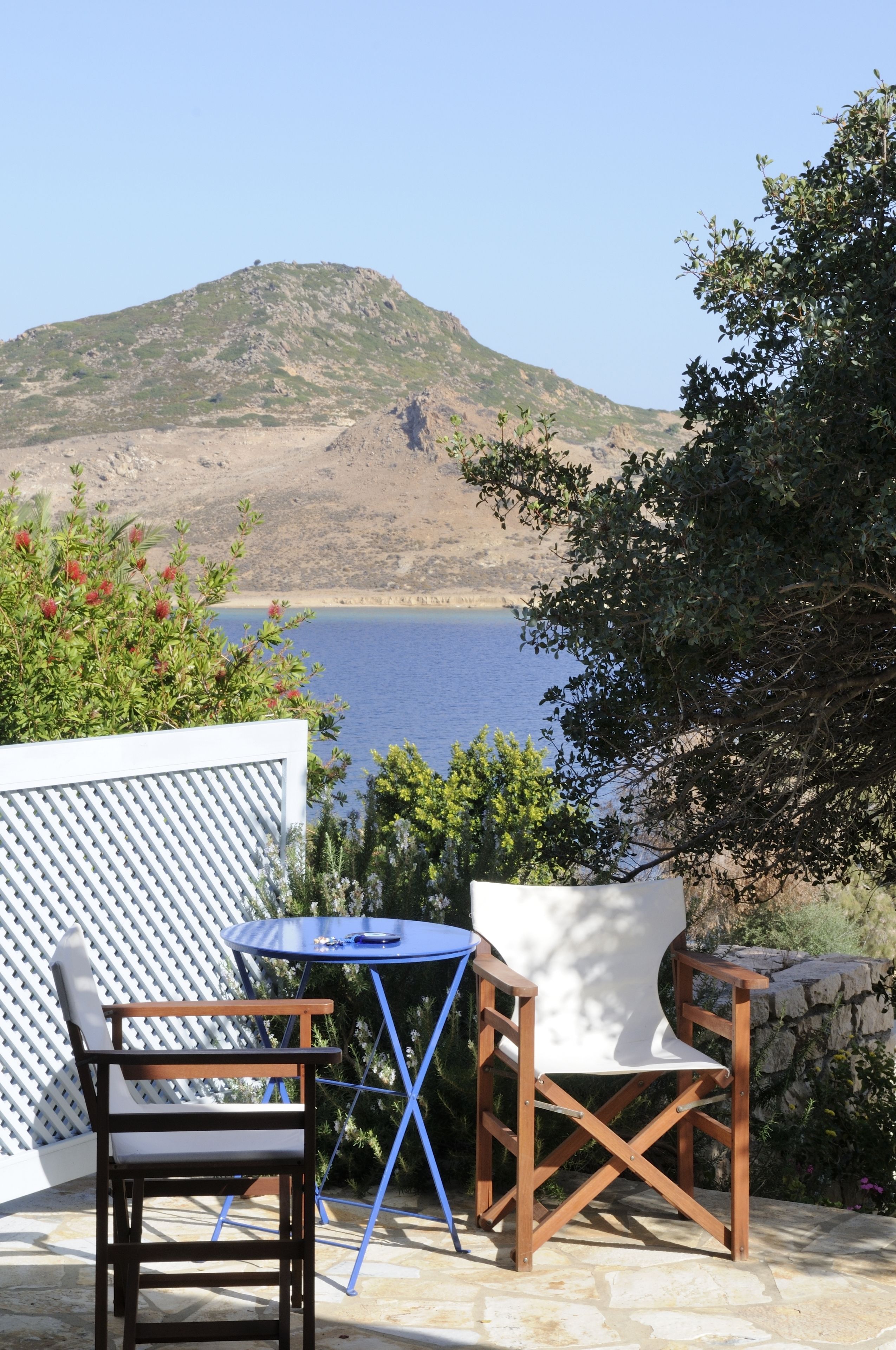 An outdoor seating area with two wooden chairs and a small table, overlooking a body of water and a hill.