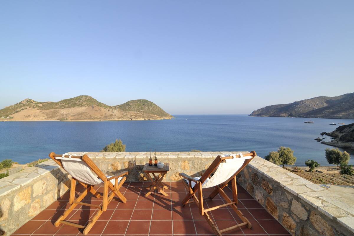 Una terraza con dos sillas de madera y una mesa, con vistas al mar tranquilo y colinas distantes.