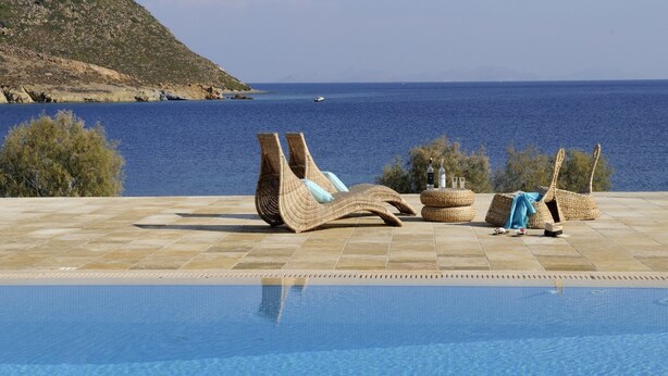 A poolside area with wicker lounge chairs and a small table with bottles, overlooking a clear blue sea and a hilly landscape. - Onar (Patmos)