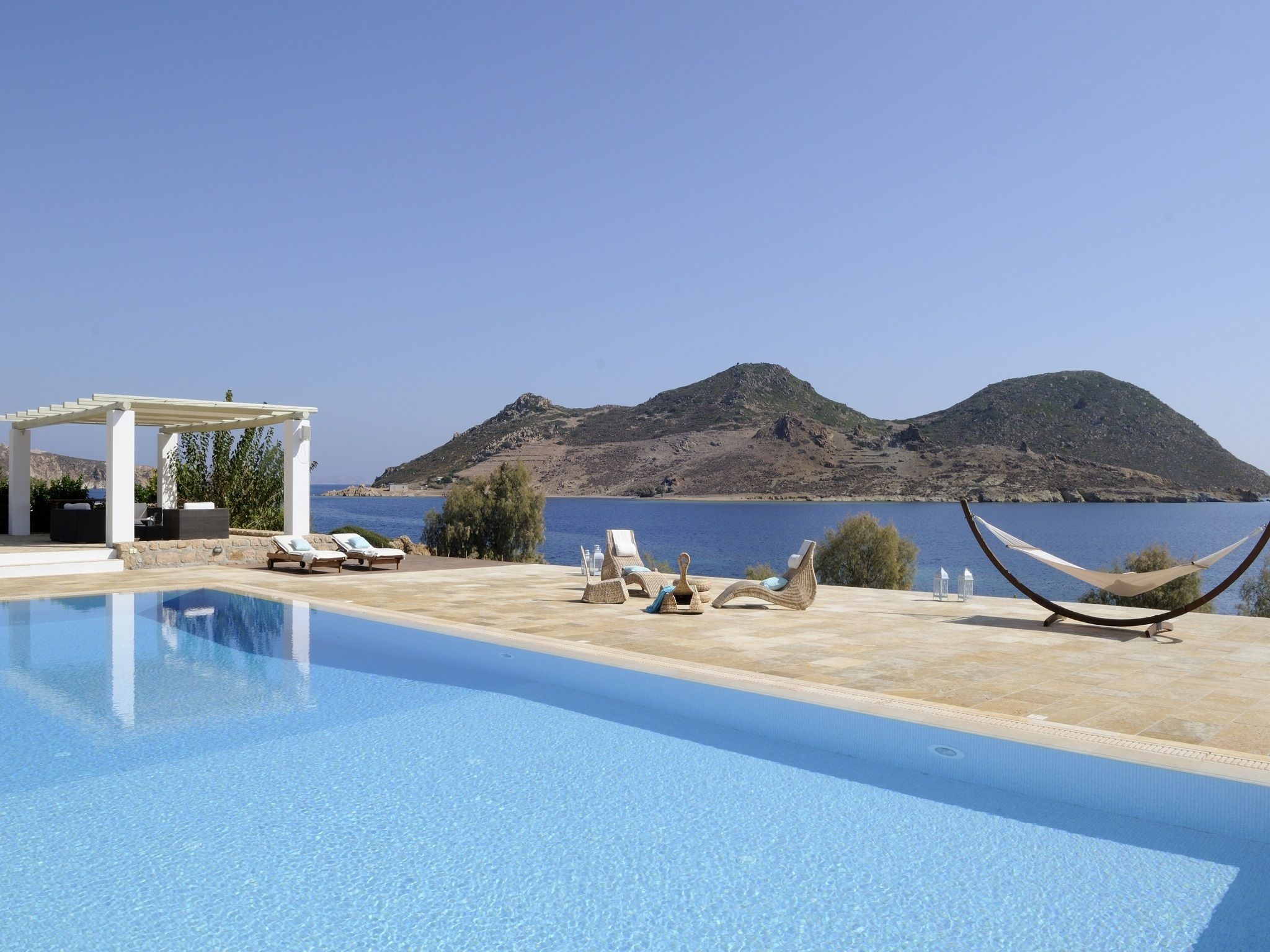 A pool area with lounge chairs and a hammock, overlooking a body of water and a mountainous landscape.