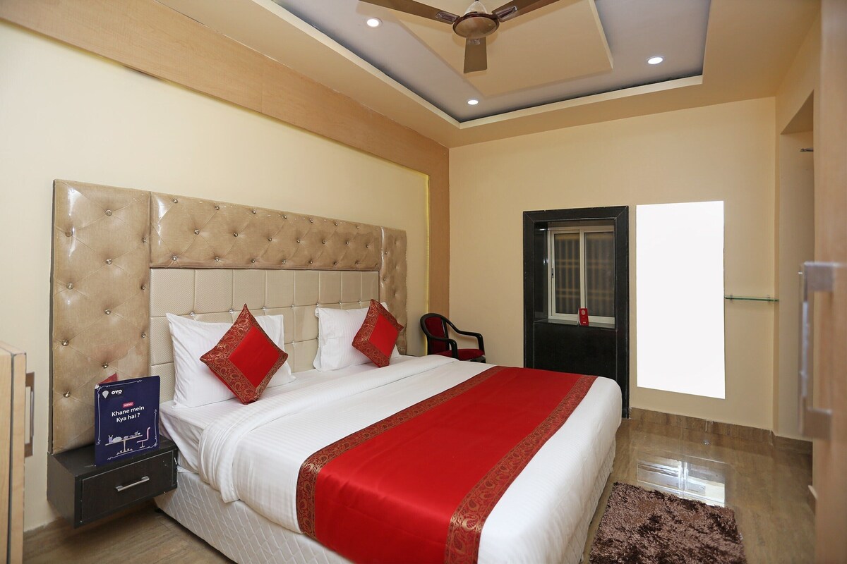 A hotel room with a tufted headboard, a large bed with white bedding and red accents, a red and gold throw blanket, a nightstand with a book, and a ceiling fan.