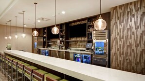 A bar area with a long counter, multiple stools, hanging pendant lights, and a modern kitchen with a refrigerator and shelves stocked with bottles. - Home2 Suites by Hilton Los Angeles Montebello (Montebello)