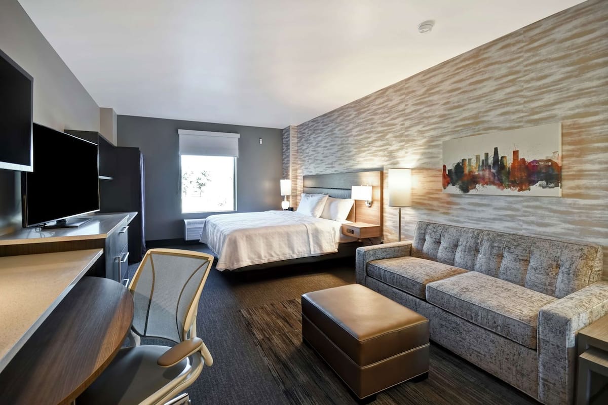 A hotel room with a bed, a sofa, a desk with a TV, a chair, and a painting on the wall.