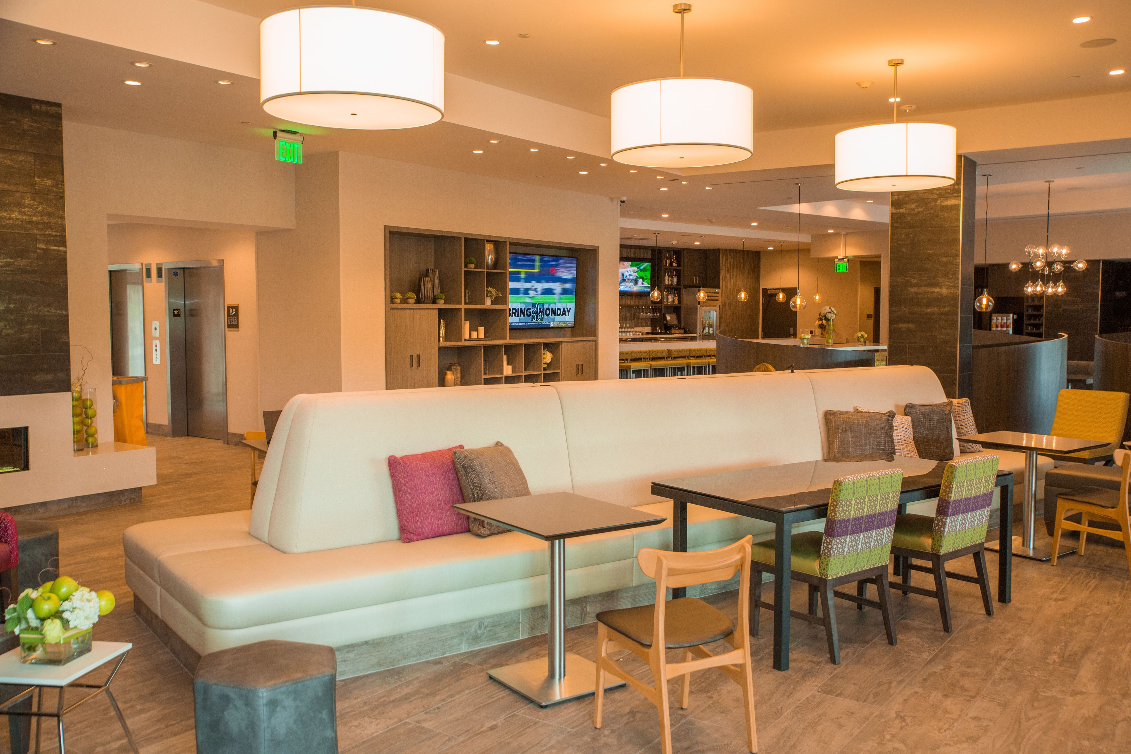 A modern hotel lobby with a seating area, a bar, and a television displaying a sports channel.
