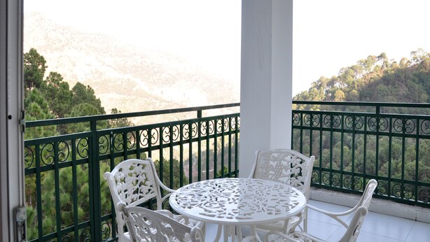 Un balcon avec une table et des chaises blanches, une rambarde en métal vert et un paysage montagneux en arrière-plan.