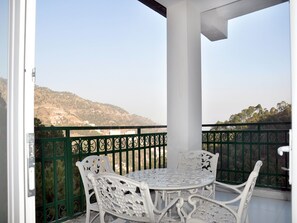 Un balcón con una mesa y sillas blancas, barandilla verde y vistas a un paisaje montañoso.