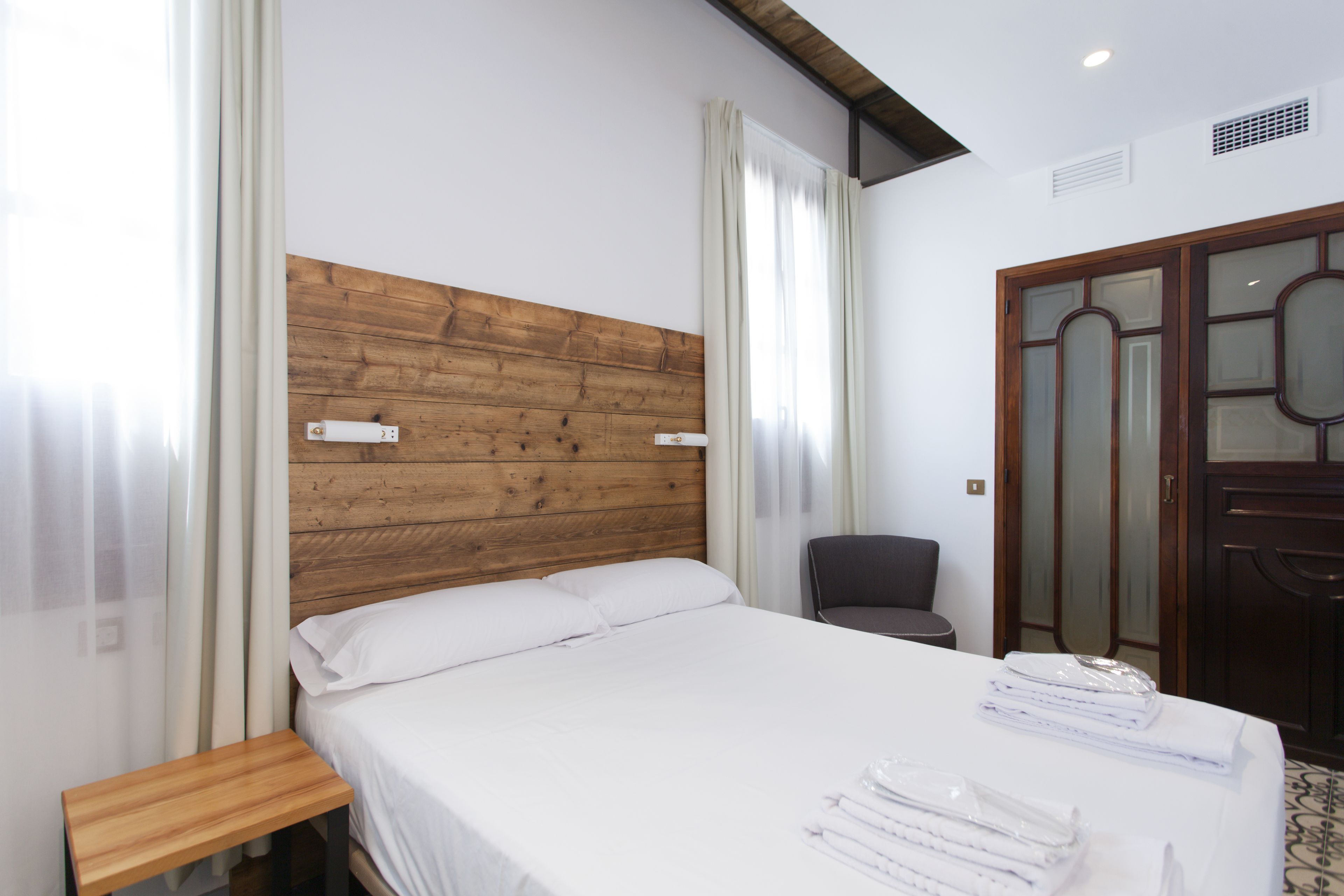 Una habitación de hotel con cabecera de madera, cama con ropa blanca, mesita de noche de madera y una silla.