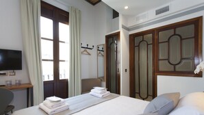 A hotel room with a neatly made bed, a desk with a flat-screen TV, a chair, and a wardrobe with a mirror.