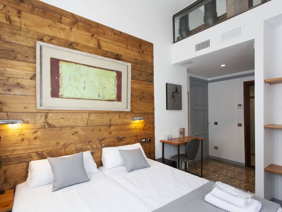 A hotel room with a wooden headboard, a large framed map, a bed with white linens, a desk with a chair, and a small plant.