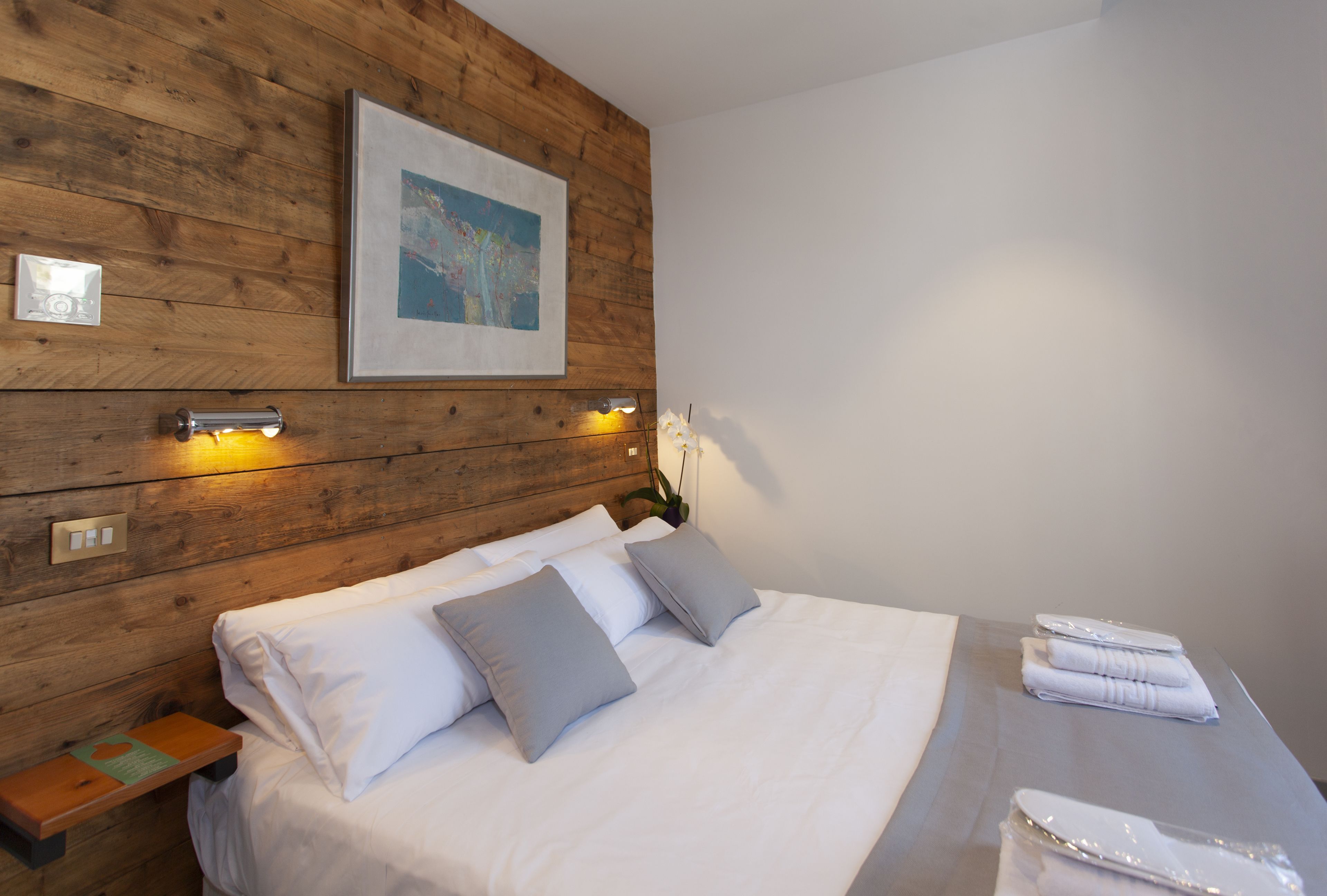 A neatly made bed with white linens and gray pillows, a wooden headboard, a framed map, and a small bedside table with a lamp.