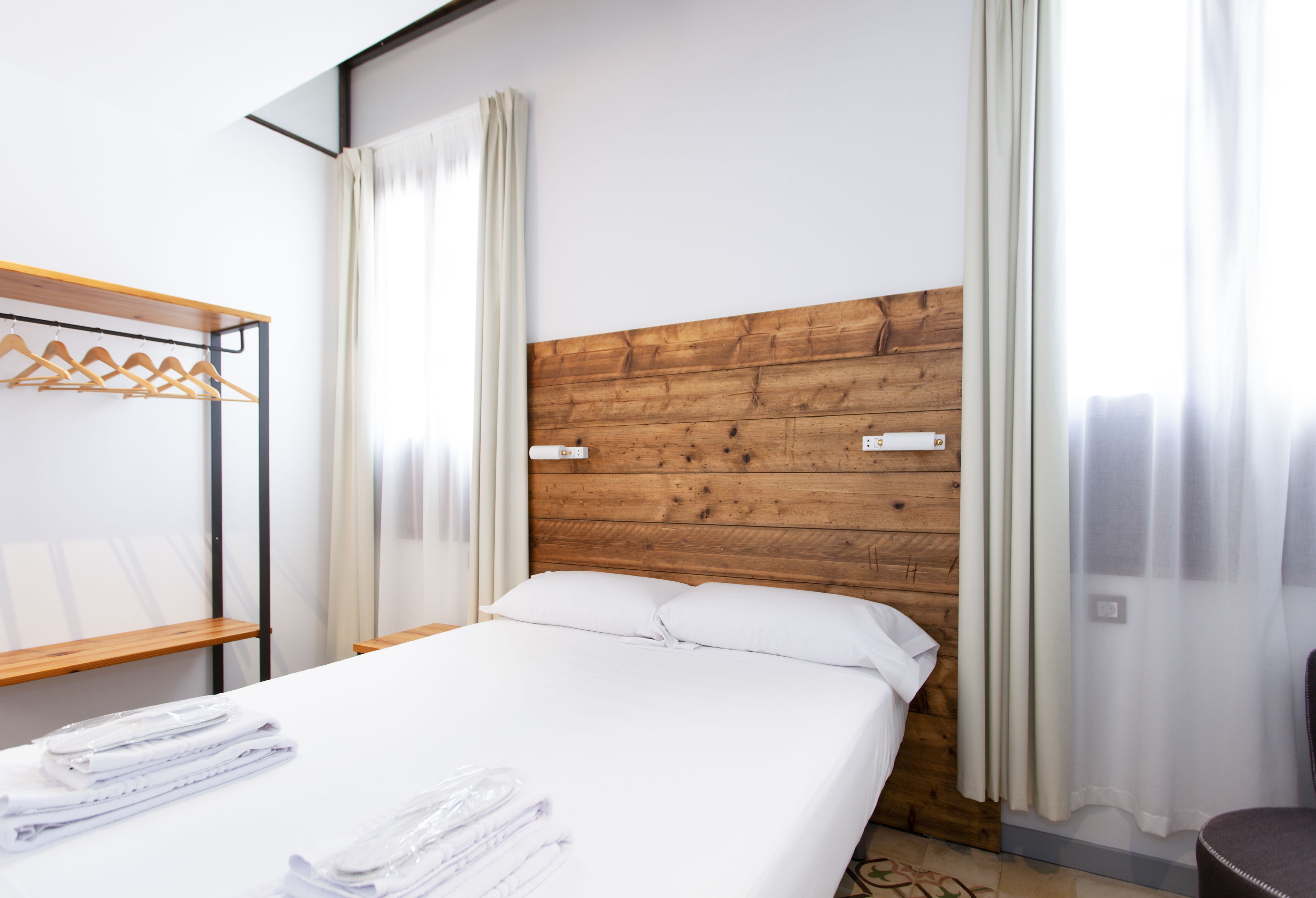 Habitación de hotel con cabecera de madera, ropa de cama blanca, estante de madera y ventana con cortinas.