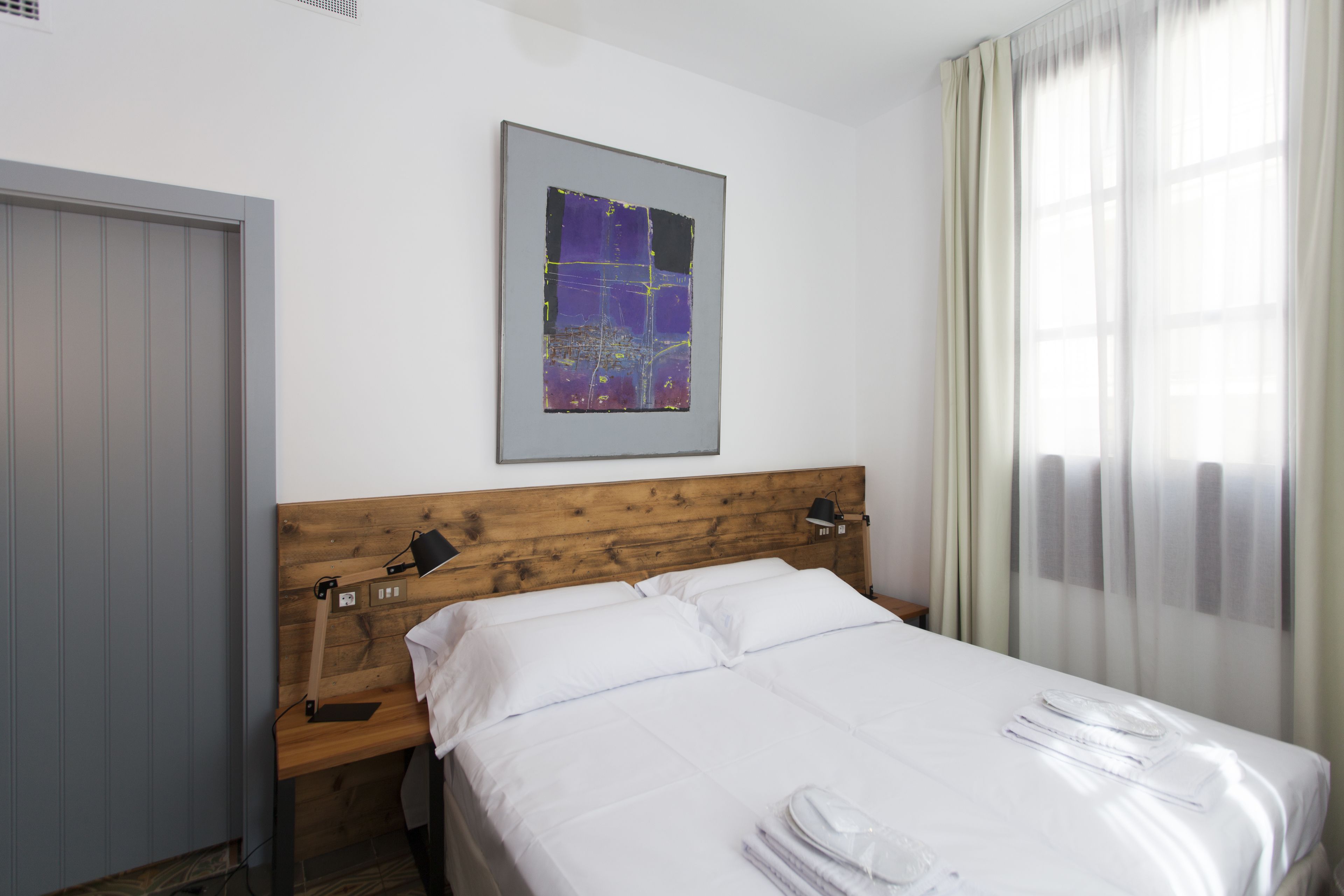 A hotel room with a wooden headboard, a bed with white linens, a bedside table with a lamp, and a window with sheer curtains.