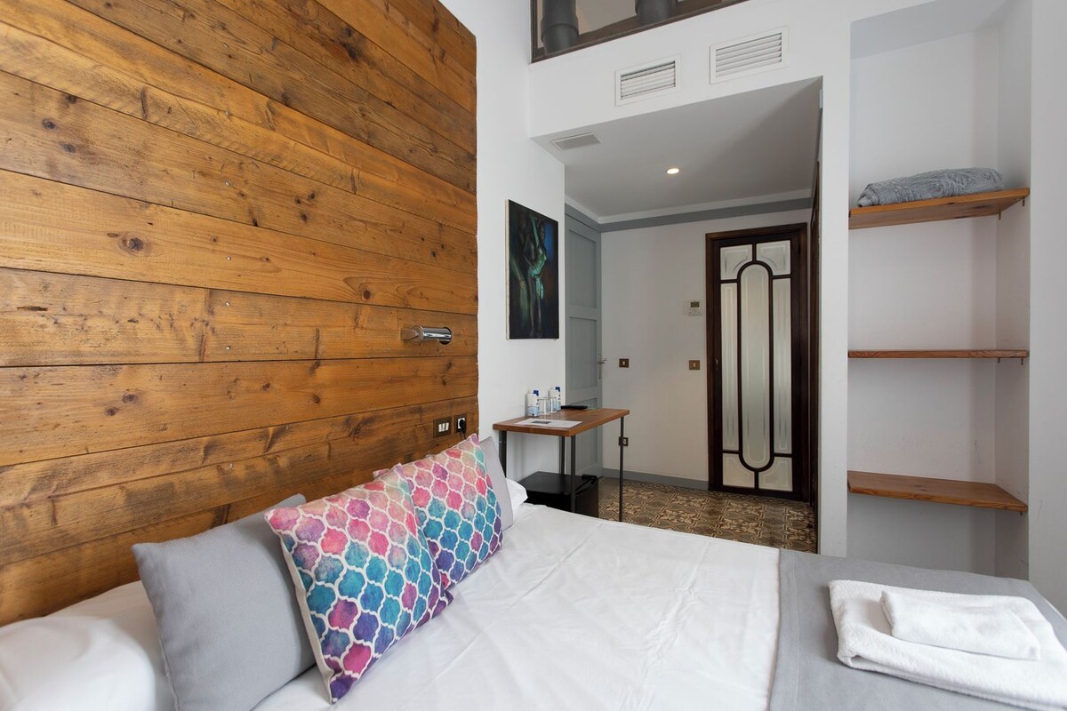Habitación de hotel con cabecera de madera, cama con ropa blanca, almohada colorida, una mesita con una botella y una puerta que da a un pasillo.