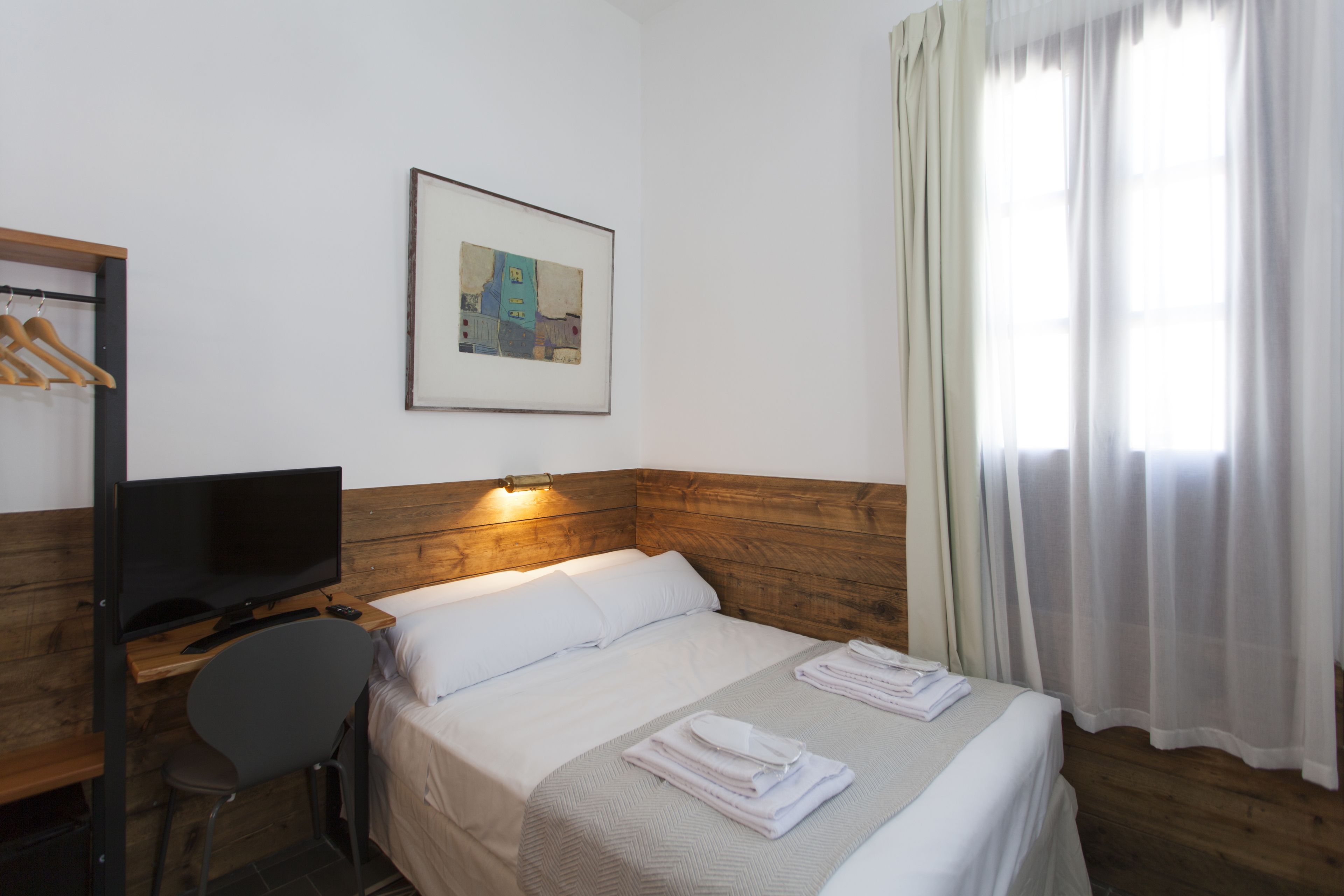 A hotel room with a wooden headboard, a bed with white linens, a television on a wooden desk, a chair, a framed picture on the wall, and a window with sheer curtains.