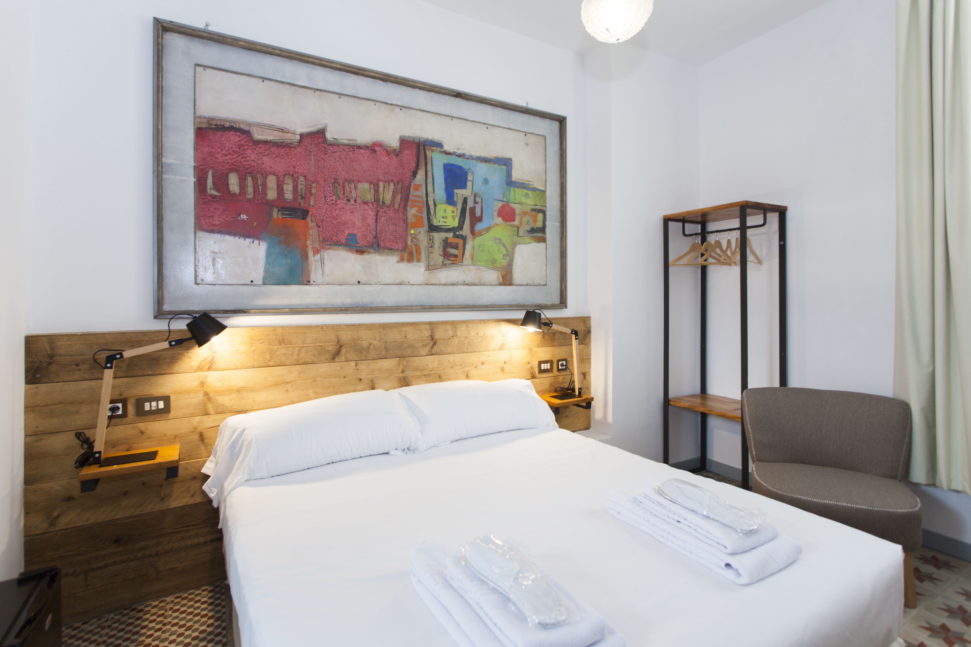 Une chambre d’hôtel avec une tête de lit en bois, un grand tableau abstrait, un lit avec du linge de maison blanc, une chaise et un porte-manteau.