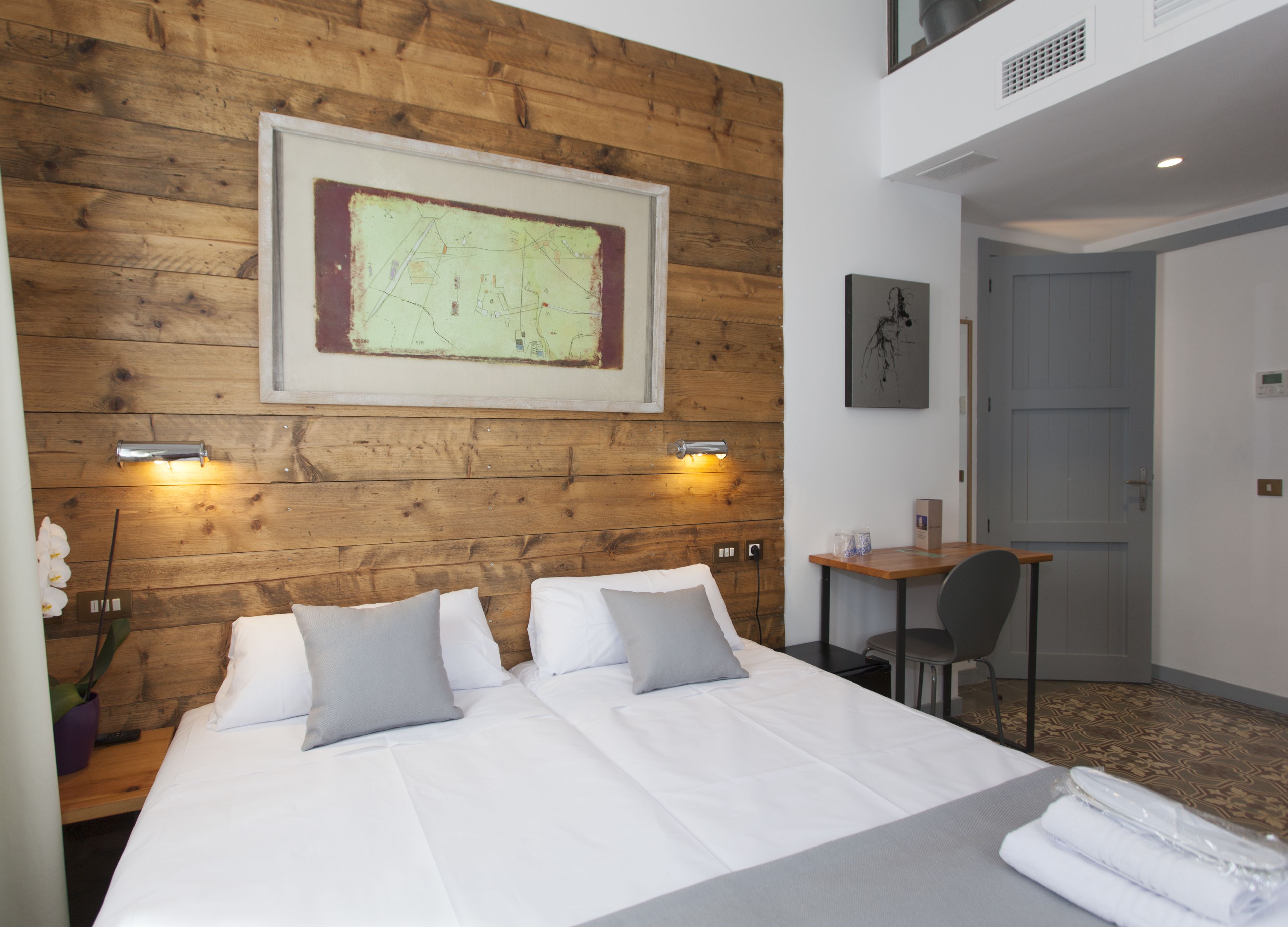 Habitación de hotel con cabecera de madera, cama grande con ropa blanca, dos mesitas de noche con lámparas, escritorio con silla y un mapa enmarcado en la pared.