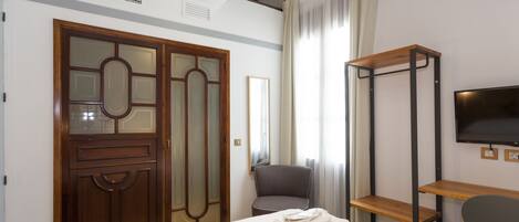 A hotel room with a bed, a chair, a TV, and a wooden shelf.