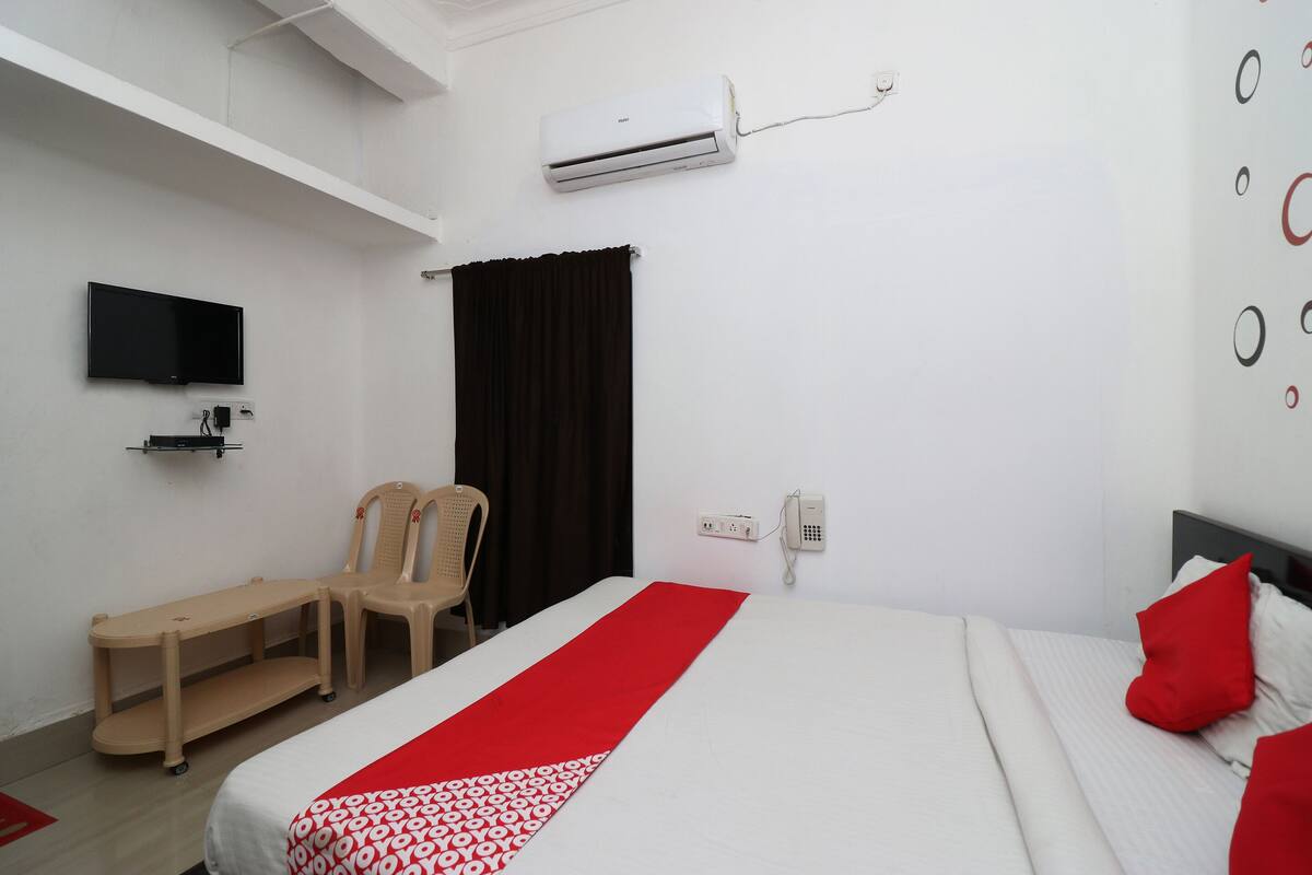 Quarto de hotel com cama, televisão fixada na parede, ar condicionado, telefone e uma cadeira.