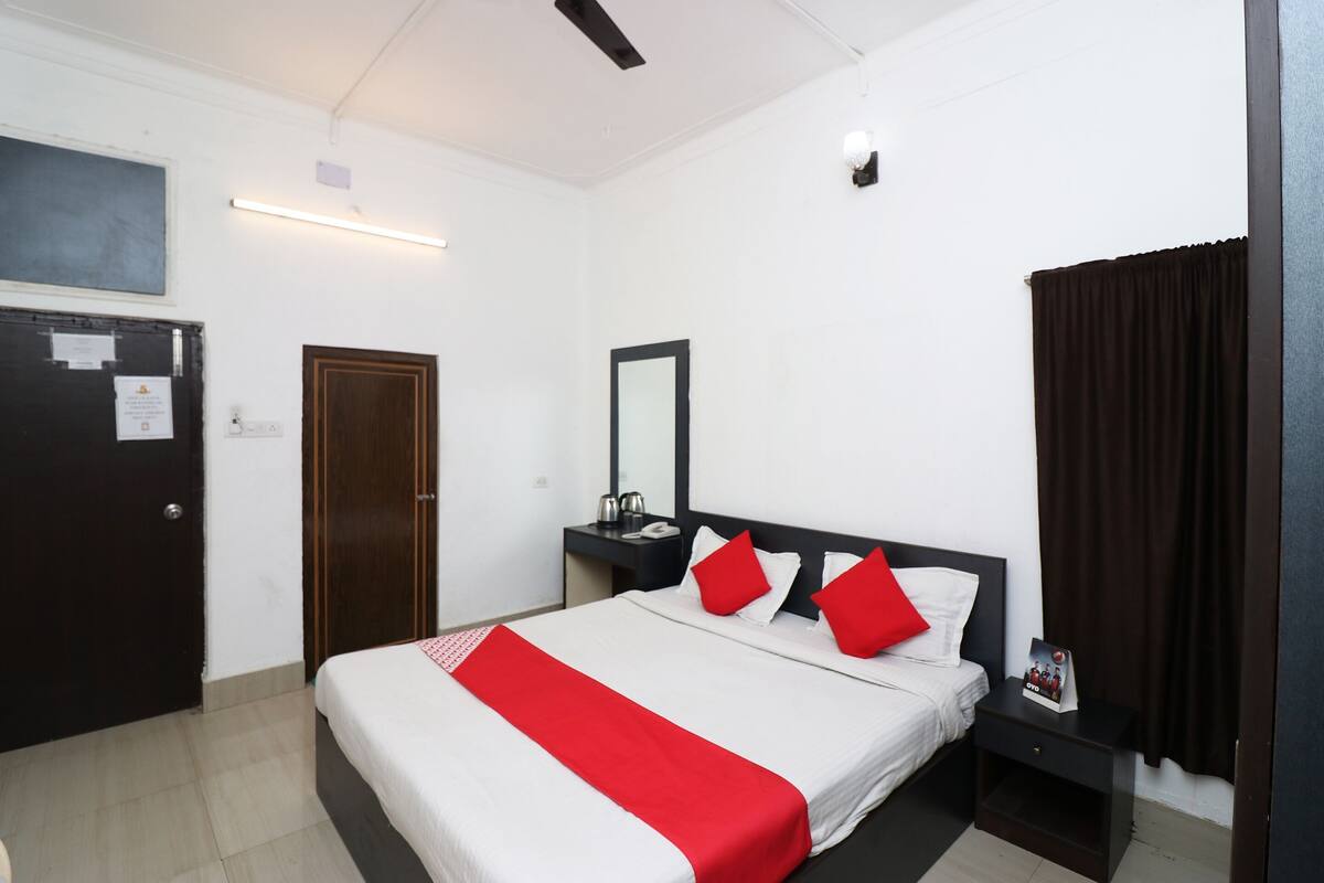 Quarto de hotel com cama, mesa de cabeceira, escrivaninha, espelho, porta e janela com cortinas.