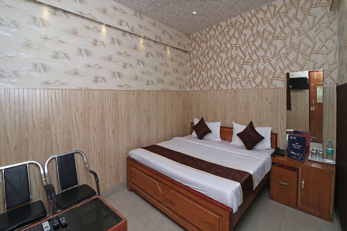 A hotel room with a wooden bed, a small desk with a chair, a mirror, and a wall with patterned wallpaper.
