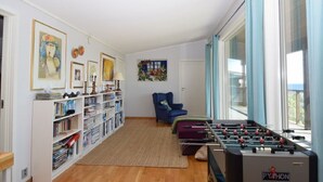 Uma sala de estar com mesa de futebol, uma poltrona azul e uma estante repleta de livros e objetos decorativos.