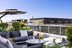 Área externa com sofás, mesa e guarda-sol, com vista para um edifício moderno com uma torre singular.
