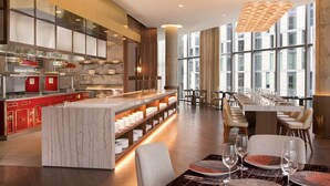 A modern hotel lobby with a marble reception desk, a long dining area with tables set for guests, and large windows offering a view of the city.