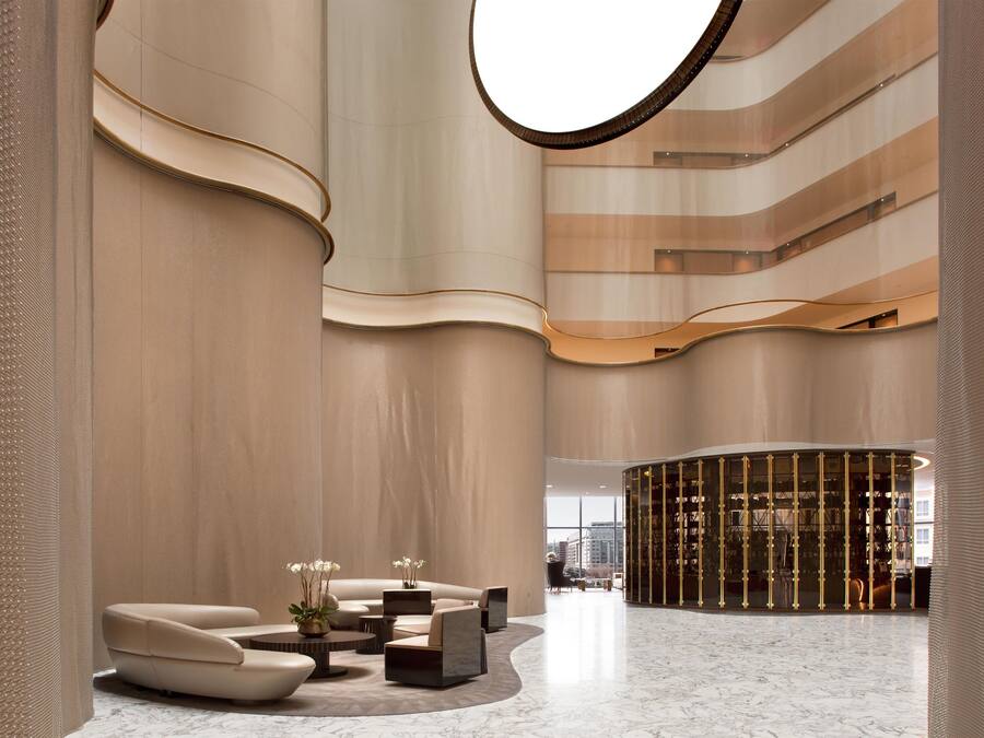 A spacious lobby with a curved seating area, a central column, and a view of the cityscape through large windows.
