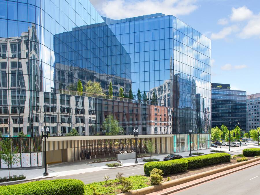 A modern glass building with a reflective facade, a landscaped area with trimmed bushes, and a clear sky.