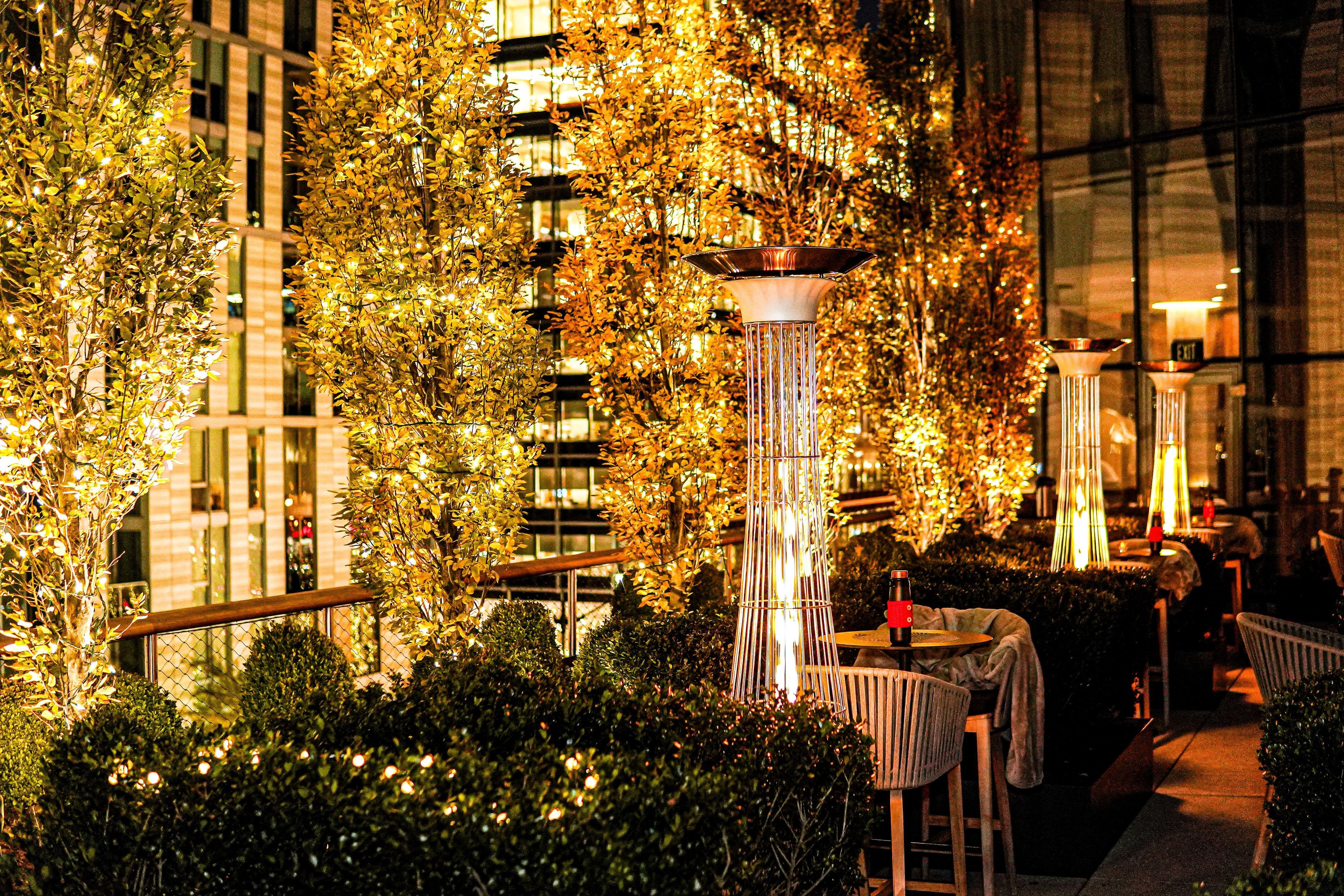 An outdoor seating area with tables and chairs, surrounded by foliage and illuminated by decorative lights.