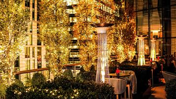 An outdoor seating area with tables and chairs, surrounded by foliage and illuminated by decorative lights.