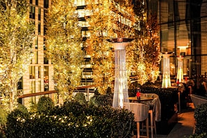 An outdoor seating area with tables and chairs, surrounded by foliage and illuminated by decorative lights. - Conrad Washington, DC (Washington)