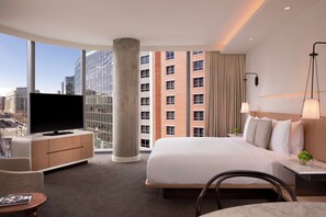 A hotel room with a large bed, a TV on a cabinet, a view of the city, and a modern lamp.