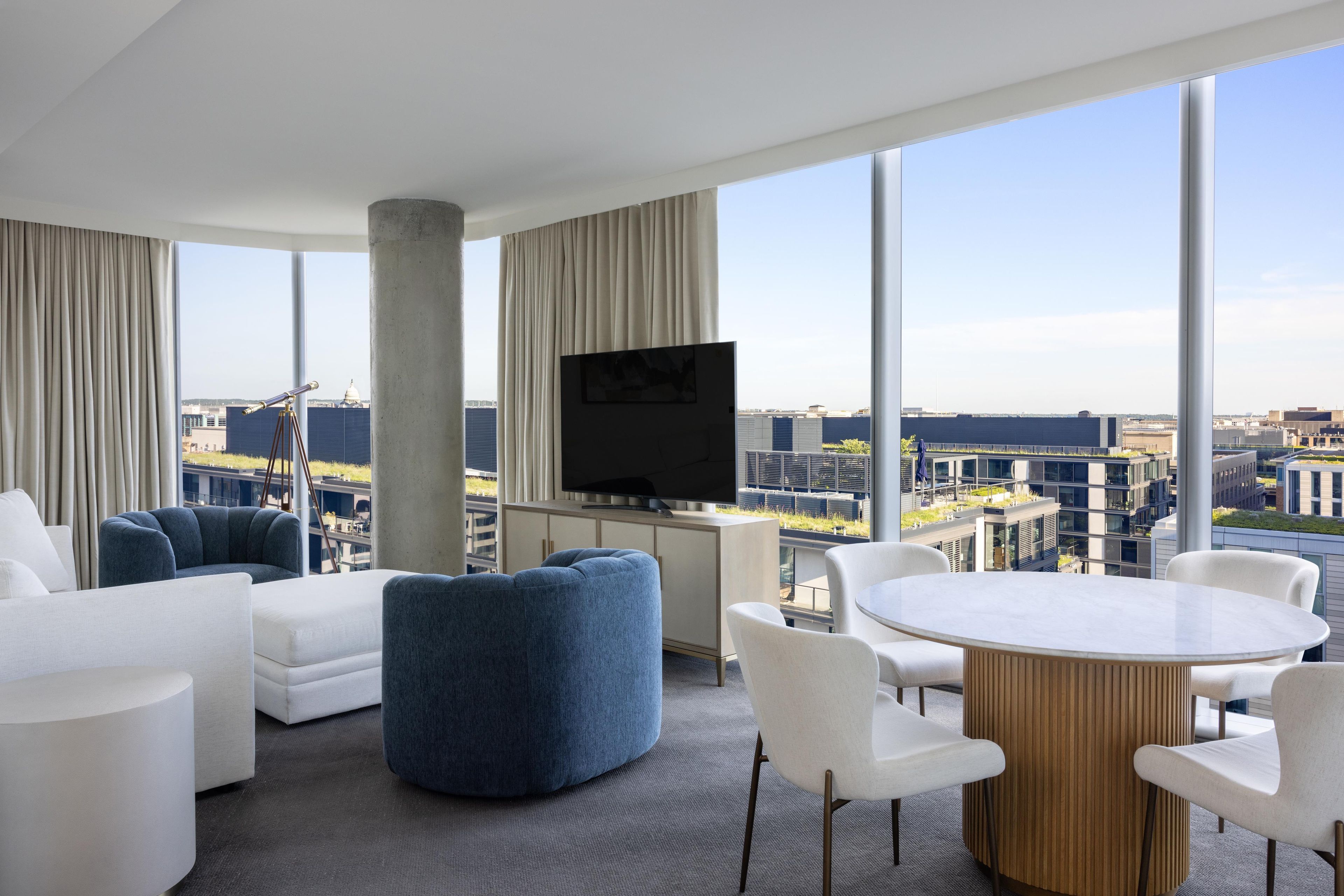 A modern hotel room with a large window, a flat-screen TV, a round table, and a sofa.